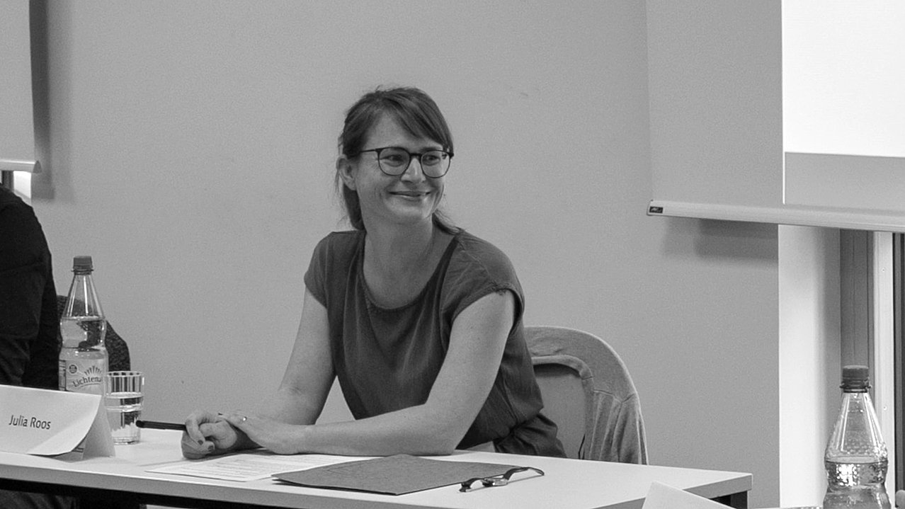 Schwarz-weiß Foto von Julia Roos, einer Frau mit Brille sitzend an einem Tisch