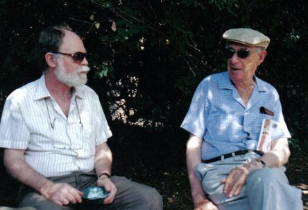 Farbfotografie zweier Männer im Freien sitzend, im Hintergrund eine Ecke. Beide Männer tragen Sonnenbrille und Hemd. Der rechte Mann hat eine Mütze auf dem Kopf und schaut den linken an, der ca. einen halben Meter von ihm entfernt sitzt und nach vorne links blickt.